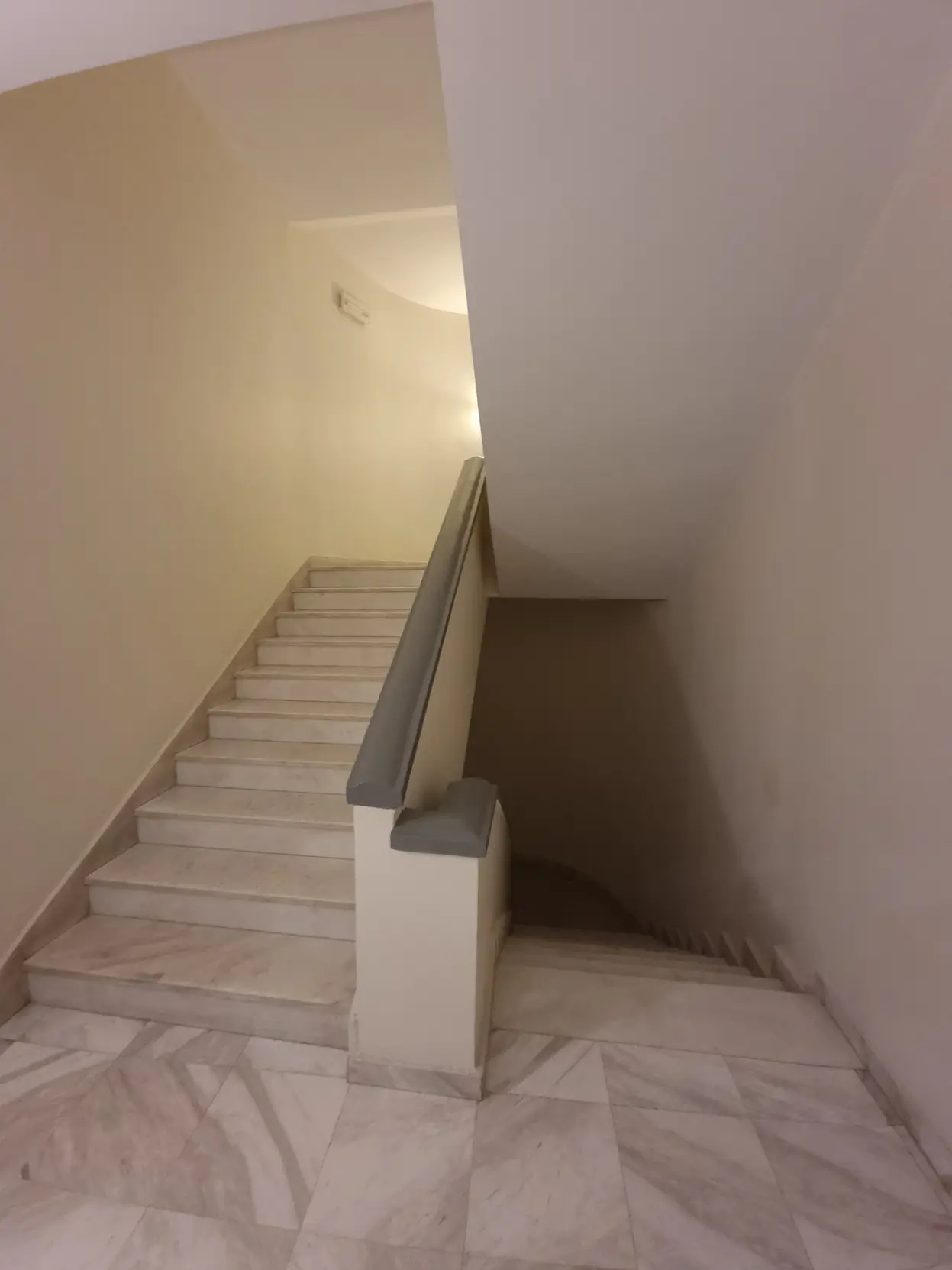 A staircase with white walls and marble floor and steps. The stairs leading down are entirely unlit, while the ones leading up are fine, giving it that subtly unsettling feeling of typical liminal scenes.