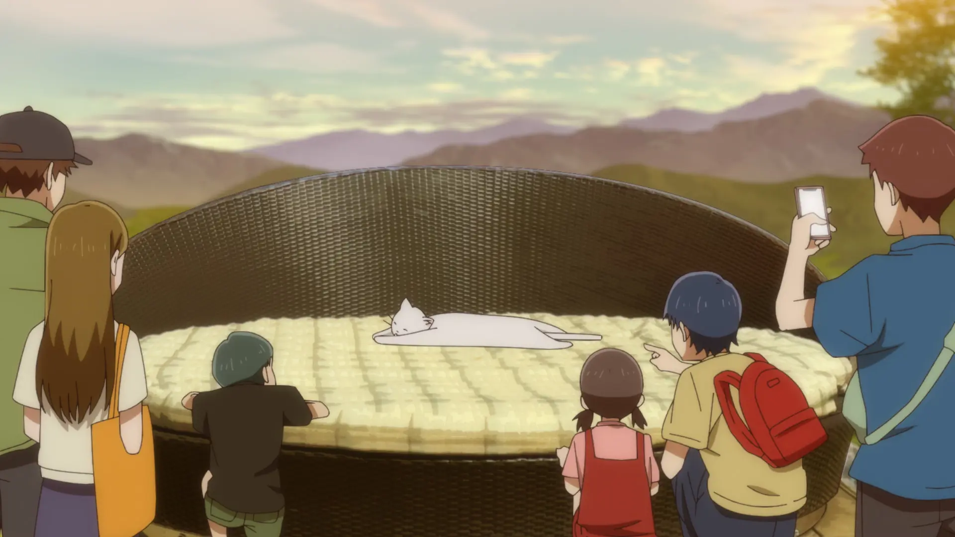 Screenshot from the anime "Mono" showing a cat snoozing on a huge comfy chair outside. There's a crowd looking at it and taking photos.